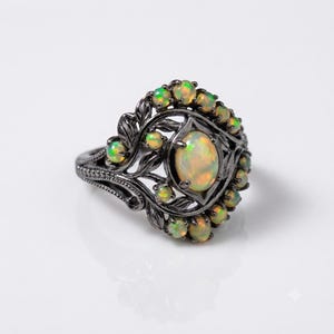 May include: A dark gunmetal ring featuring a central oval-shaped iridescent opal surrounded by smaller round opals. The band and setting are crafted with intricate leaf-like metalwork details that frame the stones in a decorative arrangement.