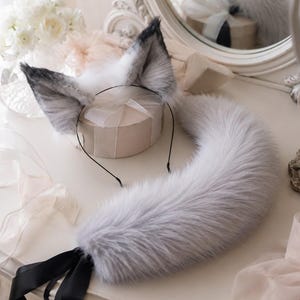 Puede incluir: Un conjunto que consta de una diadema negra fina con dos orejas de animal de piel sintética y una cola larga a juego de piel sintética. Las orejas y la cola son de color gris claro con detalles en blanco y puntas de color gris oscuro.