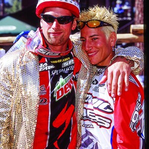 May include: Two people wearing sunglasses and colorful outfits pose for a photo. One person wears a red and black racing jersey with a Mountain Dew logo and a gold sequined jacket. The other person wears a red and white racing jersey.