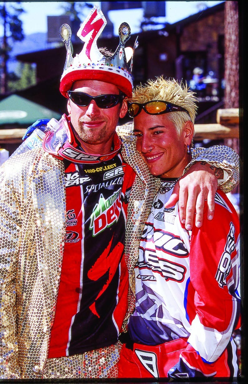 May include: Two people wearing sunglasses and colorful outfits pose for a photo. One person wears a red and black racing jersey with a Mountain Dew logo and a gold sequined jacket. The other person wears a red and white racing jersey.