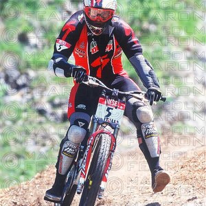 May include: A person riding a mountain bike downhill. The rider wears a red and black racing suit, a white helmet with red accents, and protective gear. The bike has a red and white frame with the word "ITALIA" on the front.