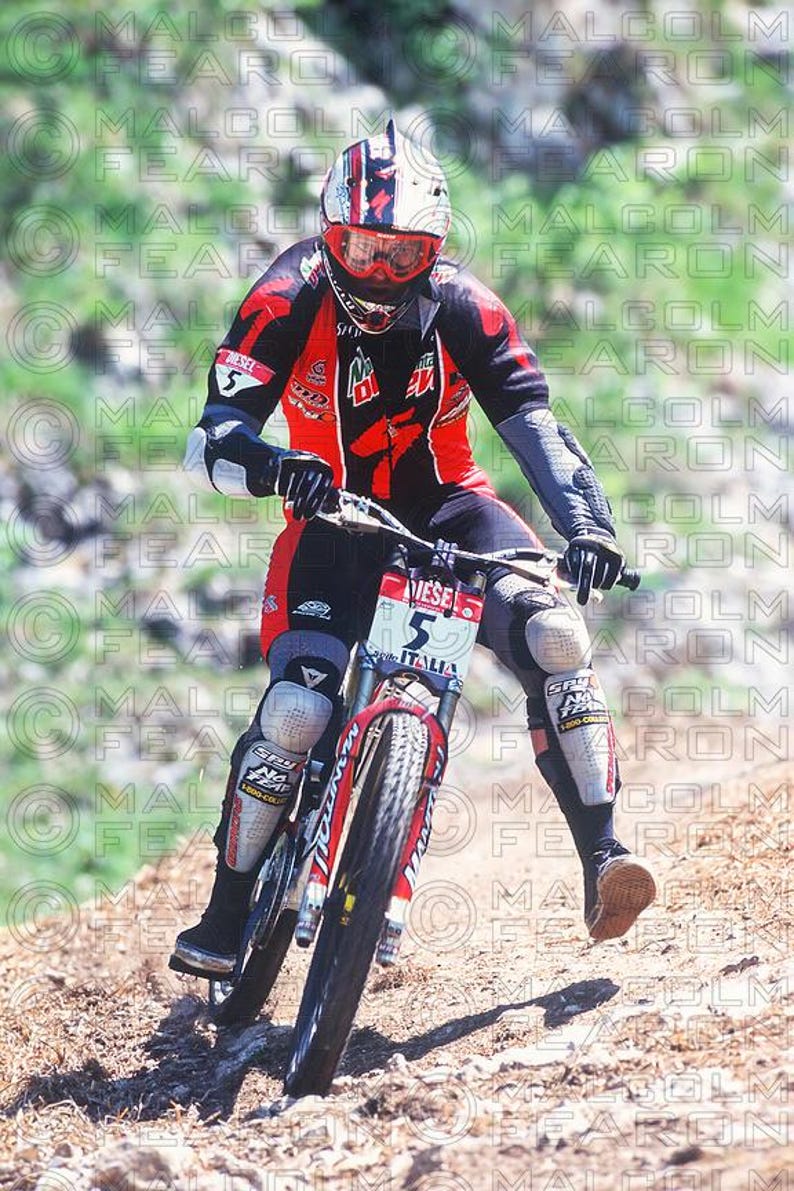 May include: A person riding a mountain bike downhill. The rider wears a red and black racing suit, a white helmet with red accents, and protective gear. The bike has a red and white frame with the word "ITALIA" on the front.
