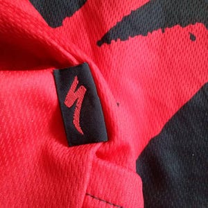 May include: Close-up of a red and black athletic jersey. The jersey has a red background with black abstract shapes. A black tag with a red logo is sewn on the jersey. The fabric has a textured, breathable design.
