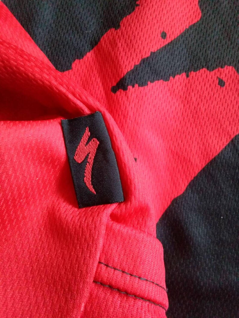 May include: Close-up of a red and black athletic jersey. The jersey has a red background with black abstract shapes. A black tag with a red logo is sewn on the jersey. The fabric has a textured, breathable design.