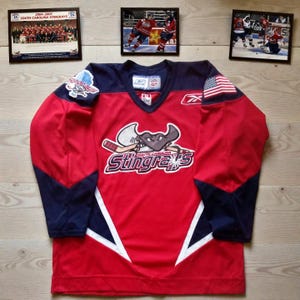 May include: A red hockey jersey with navy blue sleeves and accents. The jersey features the South Carolina Stingrays logo on the front, an American flag patch, and the Reebok logo. Framed photos of hockey players and team photos are in the background.