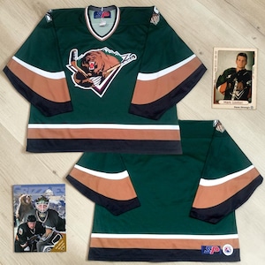 May include: A dark green hockey jersey featuring a central graphic of a brown bear holding a hockey stick. The jersey has tan and black horizontal stripes on the sleeves and hem. Included are two trading cards showing a player in the same jersey.
