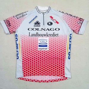 May include: A white and red short-sleeved cycling jersey with a half-zip collar. The garment features a red honeycomb pattern on the sleeves and lower torso, with printed logos including Colnago, Landbouwkrediet, Olympus, and Esprinet.