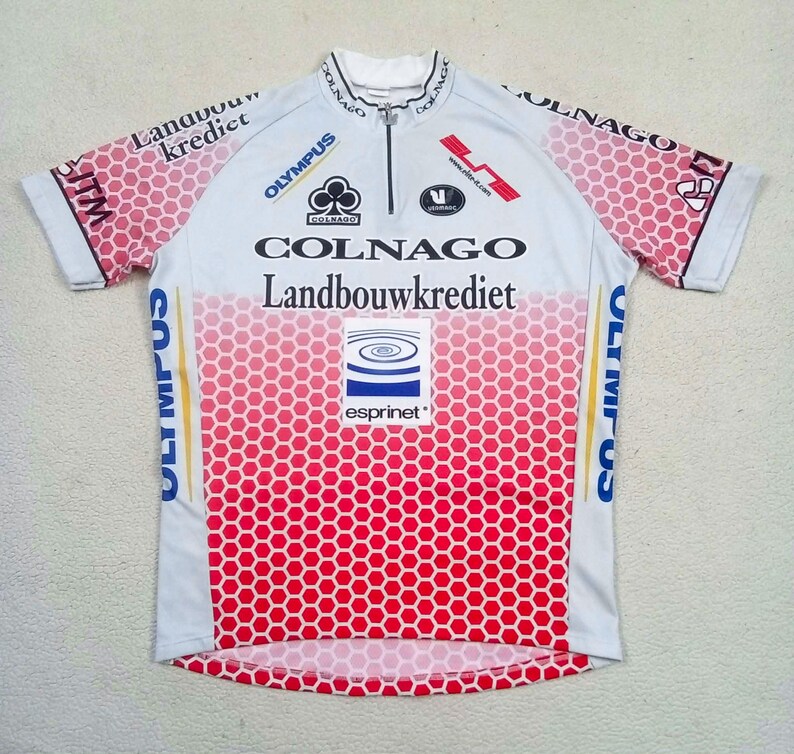 May include: A white and red short-sleeved cycling jersey with a half-zip collar. The garment features a red honeycomb pattern on the sleeves and lower torso, with printed logos including Colnago, Landbouwkrediet, Olympus, and Esprinet.