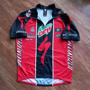 May include: A red and black Specialized cycling jersey with a half-zip collar. The jersey features the Mountain Dew logo, Specialized branding, and other sponsor logos. The sleeves are black, and the jersey has white vertical stripes down the front.