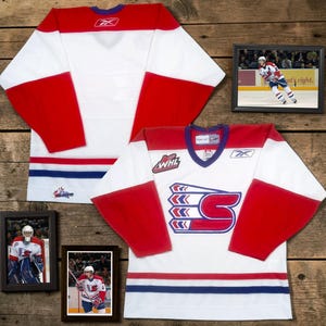 May include: A white hockey jersey with red sleeves and blue and red stripes at the hem. The front features a large stylized letter S logo and a WHL patch. The Reebok logo is on the chest and back neck. Includes two framed photos of a hockey player.