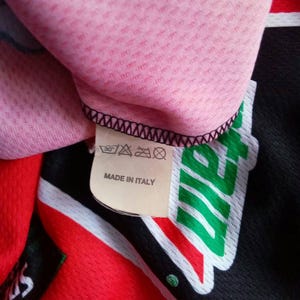 May include: Close-up of a pink, textured fabric with a black stitched edge. A tag reads "MADE IN ITALY" and features washing instructions. The garment has red, black, and green accents.
