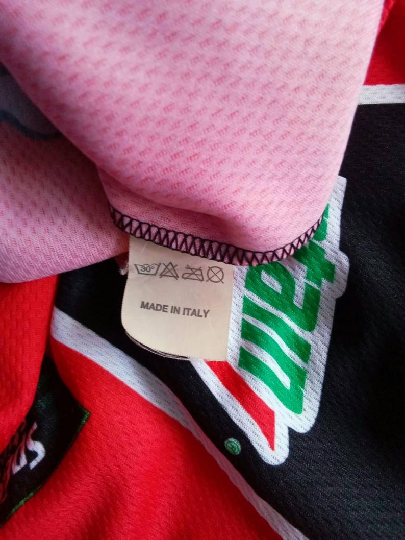 May include: Close-up of a pink, textured fabric with a black stitched edge. A tag reads "MADE IN ITALY" and features washing instructions. The garment has red, black, and green accents.