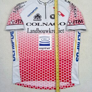 May include: A white and red cycling jersey with a honeycomb pattern, featuring logos for Colnago, Landbouwkrediet, Olympus, ITM, and Esprinet. A yellow measuring tape is placed vertically over the centre, showing a length of 68.5 cm.