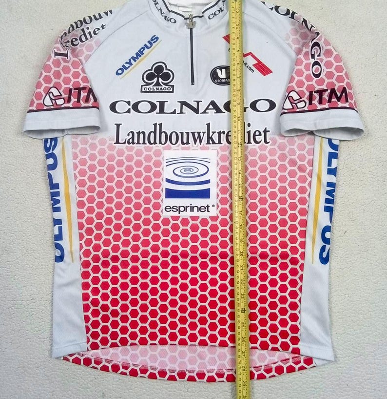 May include: A white and red cycling jersey with a honeycomb pattern, featuring logos for Colnago, Landbouwkrediet, Olympus, ITM, and Esprinet. A yellow measuring tape is placed vertically over the centre, showing a length of 68.5 cm.