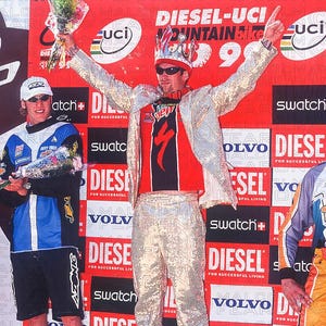 May include: A man in a sequined outfit and crown celebrates on a podium, holding flowers. He wears a red and black jersey with a logo, sunglasses, and a silver jacket. The background features event signage.