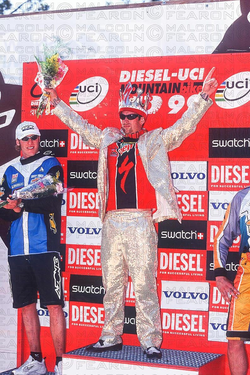 May include: A man in a sequined outfit and crown celebrates on a podium, holding flowers. He wears a red and black jersey with a logo, sunglasses, and a silver jacket. The background features event signage.