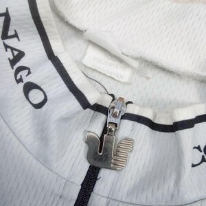 May include: A white cycling jersey with black trim and the text NAGO visible on the collar. The garment features a silver metal zipper pull shaped like a stylized bird. A white interior tag indicates the material is 100% polyester.