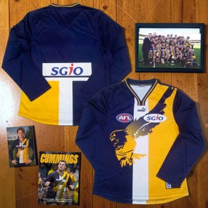 Könnte beinhalten: Ein langärmeliges West Coast Eagles AFL-Trikot in Marineblau, Gold und Weiß mit SGIO-Logo, Puma-Logo und Adlergrafik. Dazu gehören ein gerahmtes Mannschaftsfoto und zwei gedruckte Karten, die einen Spieler im gleichen Trikot zeigen.