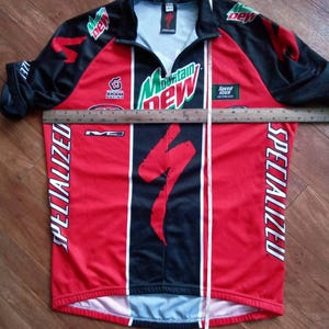 May include: A red and black Specialized cycling jersey with the Mountain Dew logo and Specialized branding. The jersey has a zipper, short sleeves, and a Specialized logo on the front. The jersey is approximately 30 inches long.