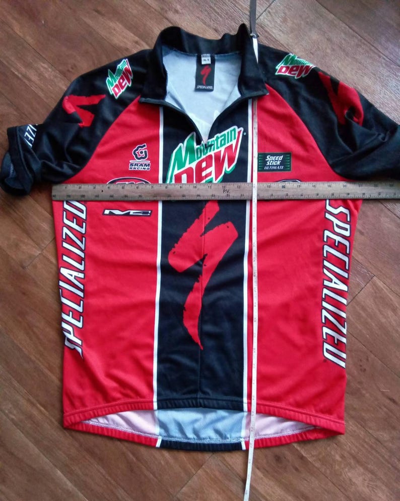 May include: A red and black Specialized cycling jersey with the Mountain Dew logo and Specialized branding. The jersey has a zipper, short sleeves, and a Specialized logo on the front. The jersey is approximately 30 inches long.
