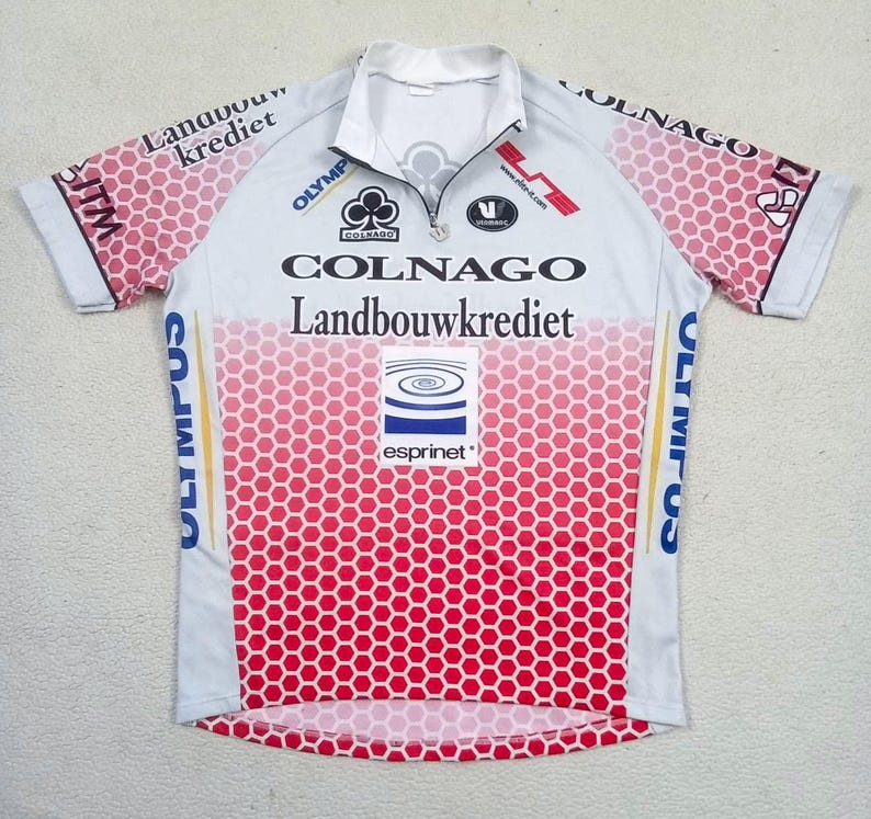 May include: A short-sleeved cycling jersey featuring a white and red honeycomb pattern. The front displays the text Colnago, Landbouwkrediet, Olympus, and Esprinet. It has a quarter-zip collar and various sponsor logos printed across the chest and sleeves.
