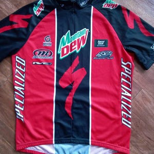 May include: A red, black, and white Specialized cycling jersey with Mountain Dew branding. The jersey features the Specialized logo, sponsor logos, and vertical white stripes. The jersey has a short zipper and short sleeves.