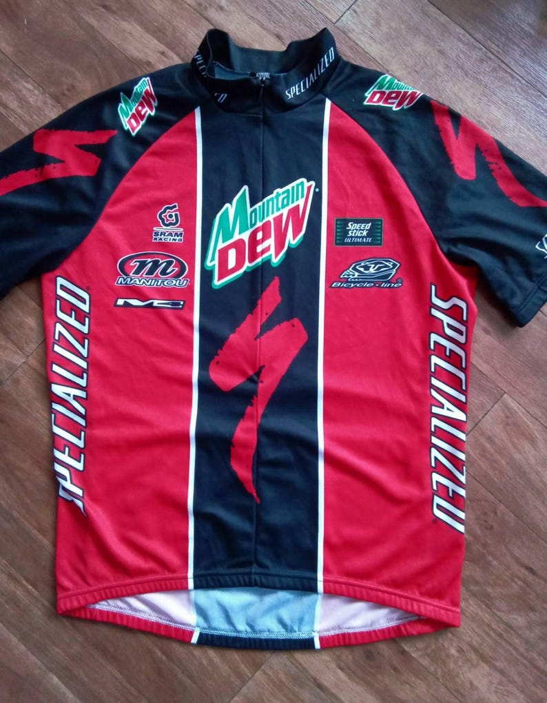 May include: A red, black, and white Specialized cycling jersey with Mountain Dew branding. The jersey features the Specialized logo, sponsor logos, and vertical white stripes. The jersey has a short zipper and short sleeves.