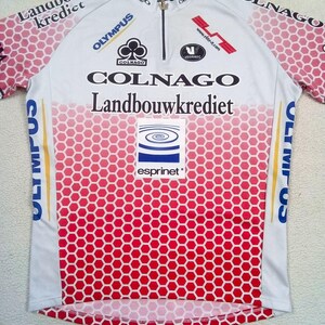 May include: A white and red cycling jersey featuring a honeycomb pattern on the lower section and sleeves. The shirt includes a front zip and various printed logos including Colnago, Landbouwkrediet, Olympus, and Esprinet. The design incorporates text and brand graphics across the chest and shoulders.