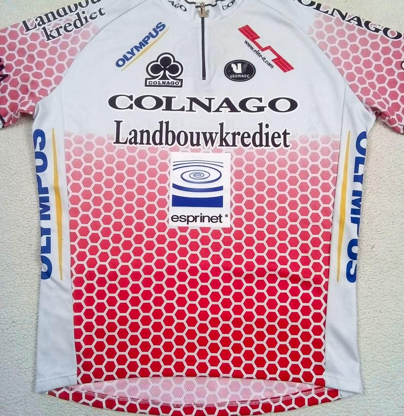 May include: A white and red cycling jersey featuring a honeycomb pattern on the lower section and sleeves. The shirt includes a front zip and various printed logos including Colnago, Landbouwkrediet, Olympus, and Esprinet. The design incorporates text and brand graphics across the chest and shoulders.