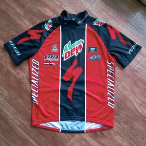 May include: A Specialized cycling jersey in red, black, and white. The jersey features the Mountain Dew logo, Specialized branding, and other sponsor logos. The jersey has a zipper and short sleeves.