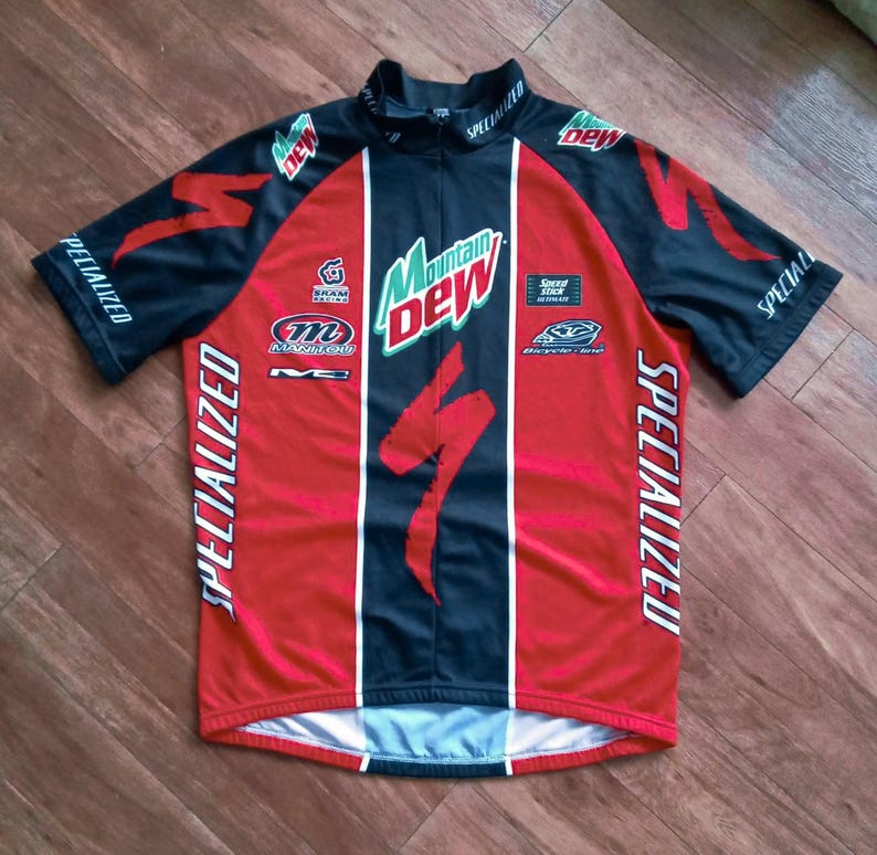 May include: A Specialized cycling jersey in red, black, and white. The jersey features the Mountain Dew logo, Specialized branding, and other sponsor logos. The jersey has a zipper and short sleeves.