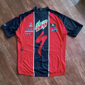 May include: Red and black cycling jersey with the Mountain Dew logo and Specialized branding. The jersey has a black collar and sleeves, with white vertical stripes and various sponsor logos.
