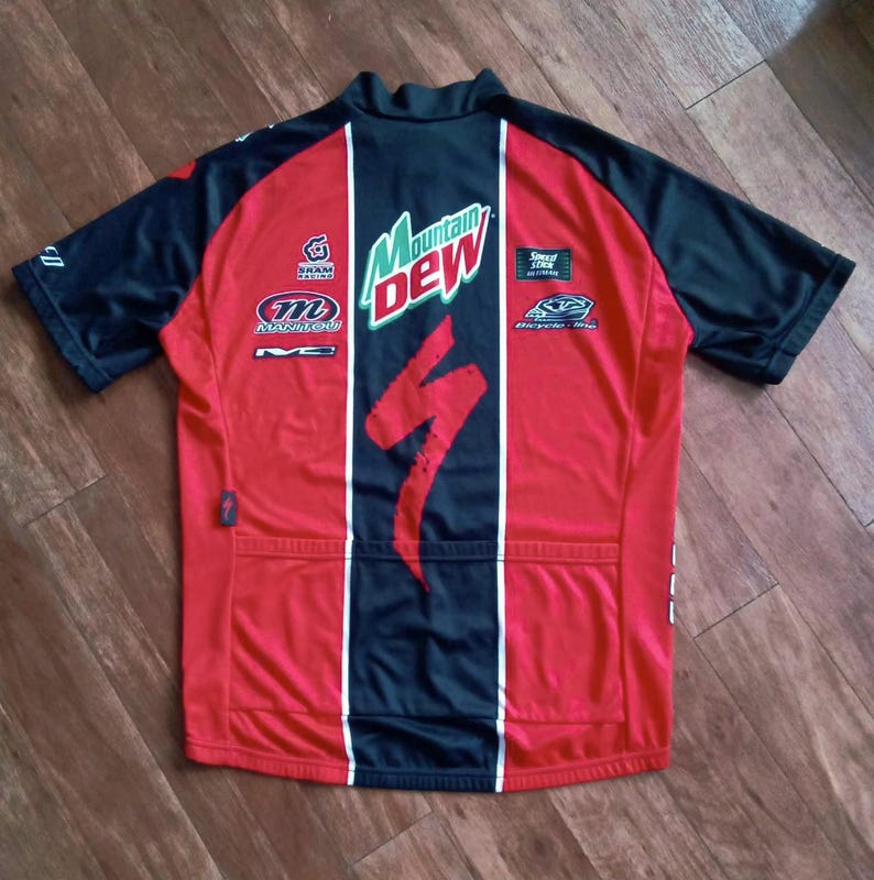 May include: Red and black cycling jersey with the Mountain Dew logo and Specialized branding. The jersey has a black collar and sleeves, with white vertical stripes and various sponsor logos.
