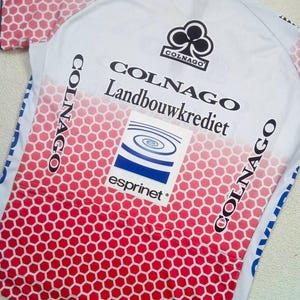 May include: A white cycling jersey featuring a red honeycomb pattern that fades towards the top. The shirt displays the black Colnago logo, the text Colnago Landbouwkrediet, and a square graphic with blue concentric circles and the word esprinet.