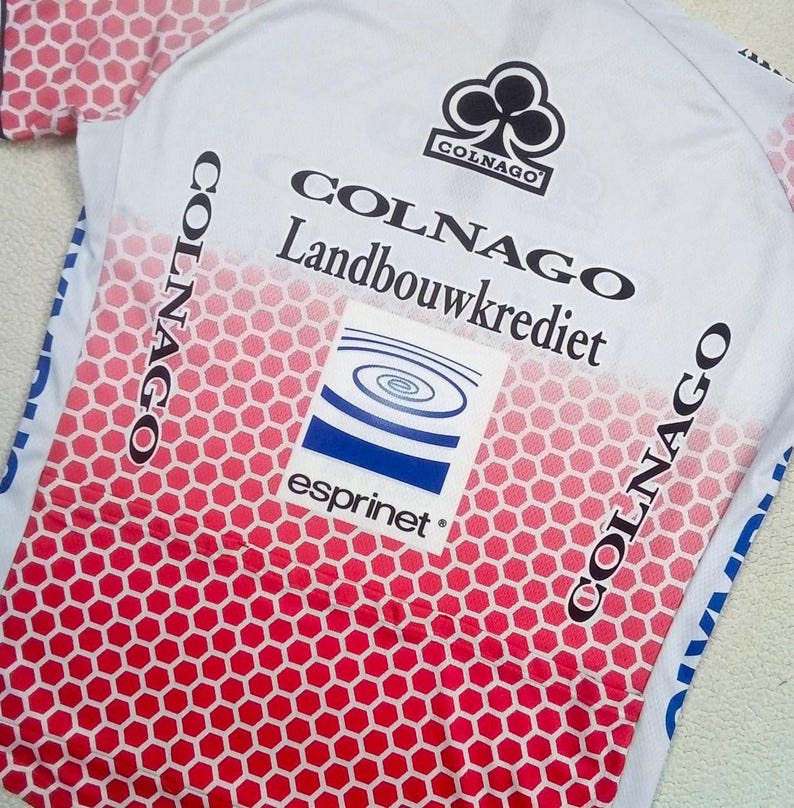 May include: A white cycling jersey featuring a red honeycomb pattern that fades towards the top. The shirt displays the black Colnago logo, the text Colnago Landbouwkrediet, and a square graphic with blue concentric circles and the word esprinet.