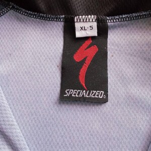 May include: Close-up of a Specialized cycling jersey tag. The tag is black with a red logo and the word "SPECIALIZED" in white. The size is XL-5. The jersey is light blue and black.