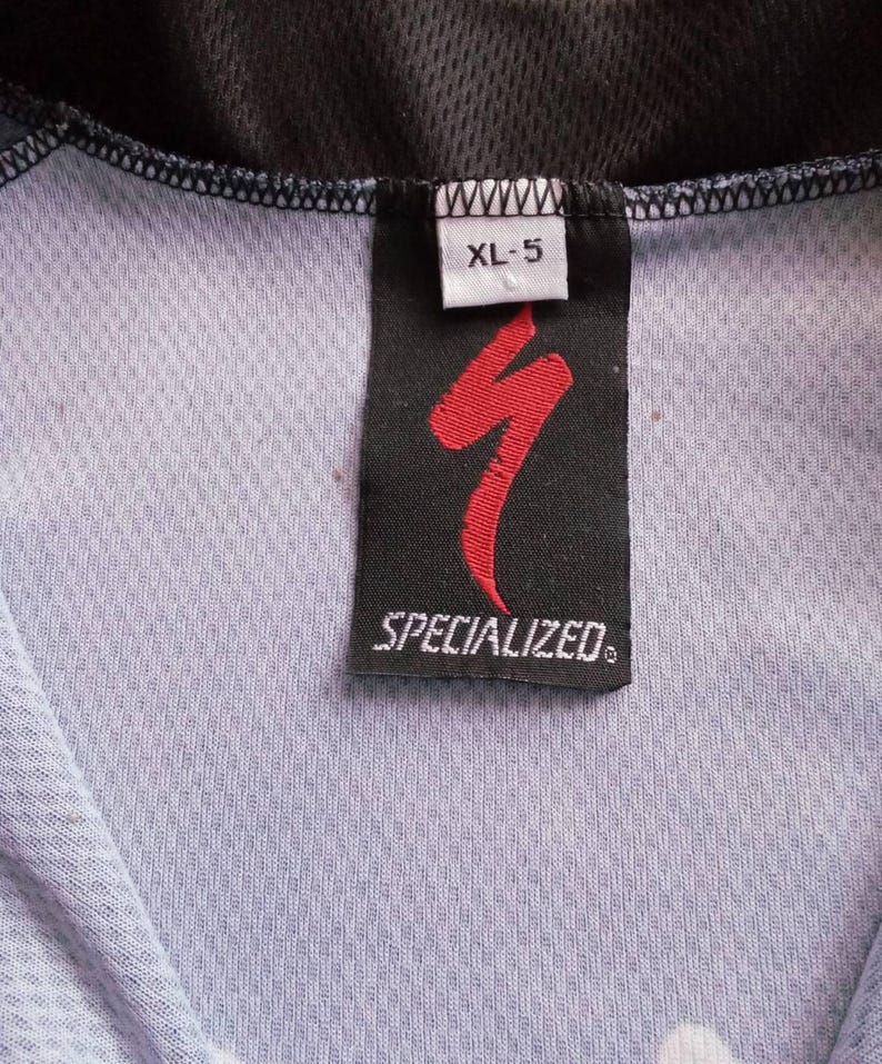 May include: Close-up of a Specialized cycling jersey tag. The tag is black with a red logo and the word "SPECIALIZED" in white. The size is XL-5. The jersey is light blue and black.