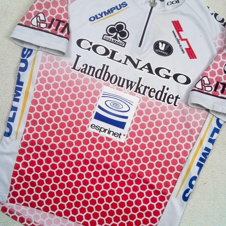 May include: A white cycling jersey with a red honeycomb pattern on the lower front and sleeves. It features logos for Colnago, Landbouwkrediet, Olympus, Elite, Vermarc, and Esprinet. The shirt has a short zip at the neck and short sleeves.