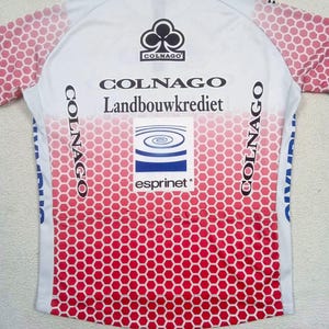 May include: A white and red cycling jersey featuring a honeycomb pattern on the sleeves and lower body. The back displays the text Colnago, Landbouwkrediet, and Esprinet, along with a Colnago logo and a blue and white spiral graphic on a square patch.