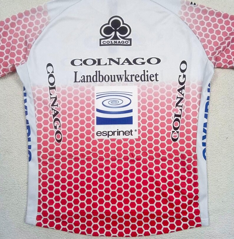 May include: A white and red cycling jersey featuring a honeycomb pattern on the sleeves and lower body. The back displays the text Colnago, Landbouwkrediet, and Esprinet, along with a Colnago logo and a blue and white spiral graphic on a square patch.