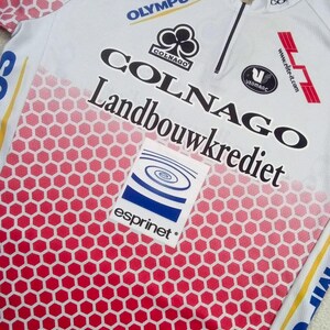 May include: A white cycling jersey with a red honeycomb pattern gradient at the bottom. The shirt features black text reading Colnago and Landbouwkrediet, with logos for Olympus, Vermarc, and Esprinet. It includes a partial front zipper.