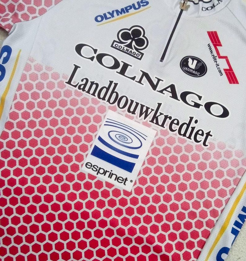 May include: A white cycling jersey with a red honeycomb pattern gradient at the bottom. The shirt features black text reading Colnago and Landbouwkrediet, with logos for Olympus, Vermarc, and Esprinet. It includes a partial front zipper.