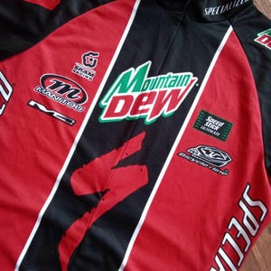 May include: A red, black, and white Specialized cycling jersey with Mountain Dew branding. The jersey features logos for SRAM Racing, Manitou, and Bicycle-line. The jersey has a Specialized logo on the sleeve and collar.