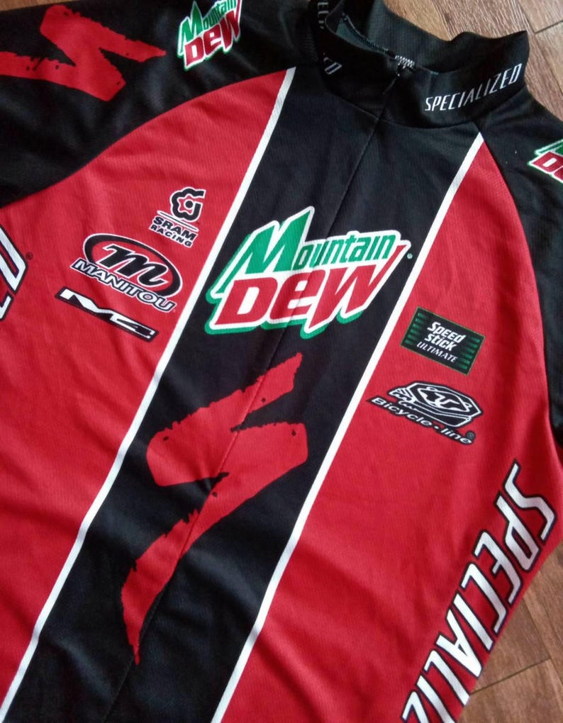 May include: A red, black, and white Specialized cycling jersey with Mountain Dew branding. The jersey features logos for SRAM Racing, Manitou, and Bicycle-line. The jersey has a Specialized logo on the sleeve and collar.
