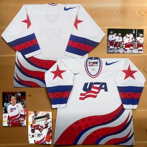 May include: A white Nike USA hockey jersey featuring red and blue wavy stripes, star patterns on the sleeves, and a large USA logo on the chest. The jersey includes a V-neck collar and is displayed alongside three photographs of players wearing the jersey.