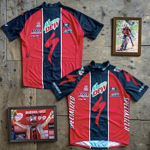 May include: Two red and black Specialized cycling jerseys with Mountain Dew logos and Specialized branding. The jerseys are displayed on a wooden surface, along with two framed photos of cycling events.