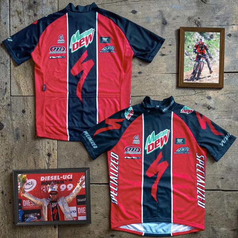 May include: Two red and black Specialized cycling jerseys with Mountain Dew logos and Specialized branding. The jerseys are displayed on a wooden surface, along with two framed photos of cycling events.