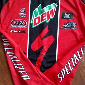 May include: Red and black long-sleeved cycling jersey with white accents. Features the "Mountain Dew" logo, "Specialized" text on the sleeves, and various sponsor logos including "SRAM Racing" and "Manitou".