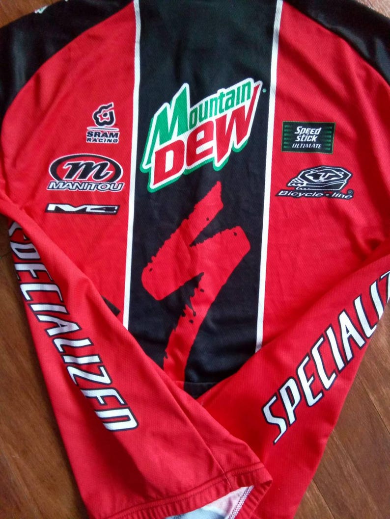 May include: Red and black long-sleeved cycling jersey with white accents. Features the "Mountain Dew" logo, "Specialized" text on the sleeves, and various sponsor logos including "SRAM Racing" and "Manitou".