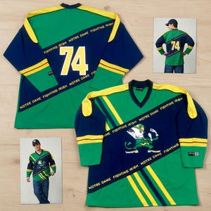 May include: Green and navy blue long-sleeved jersey with yellow accents. The jersey features the number 74 in white, along with the text "NOTRE DAME FIGHTING IRISH". A graphic of the Notre Dame leprechaun is also visible.